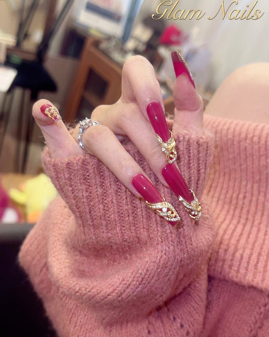 Nahaufnahme eines Daumennagels mit glänzender Magenta-Basis und einer goldfarbenen Kristallverzierung nahe der Nagelhaut.