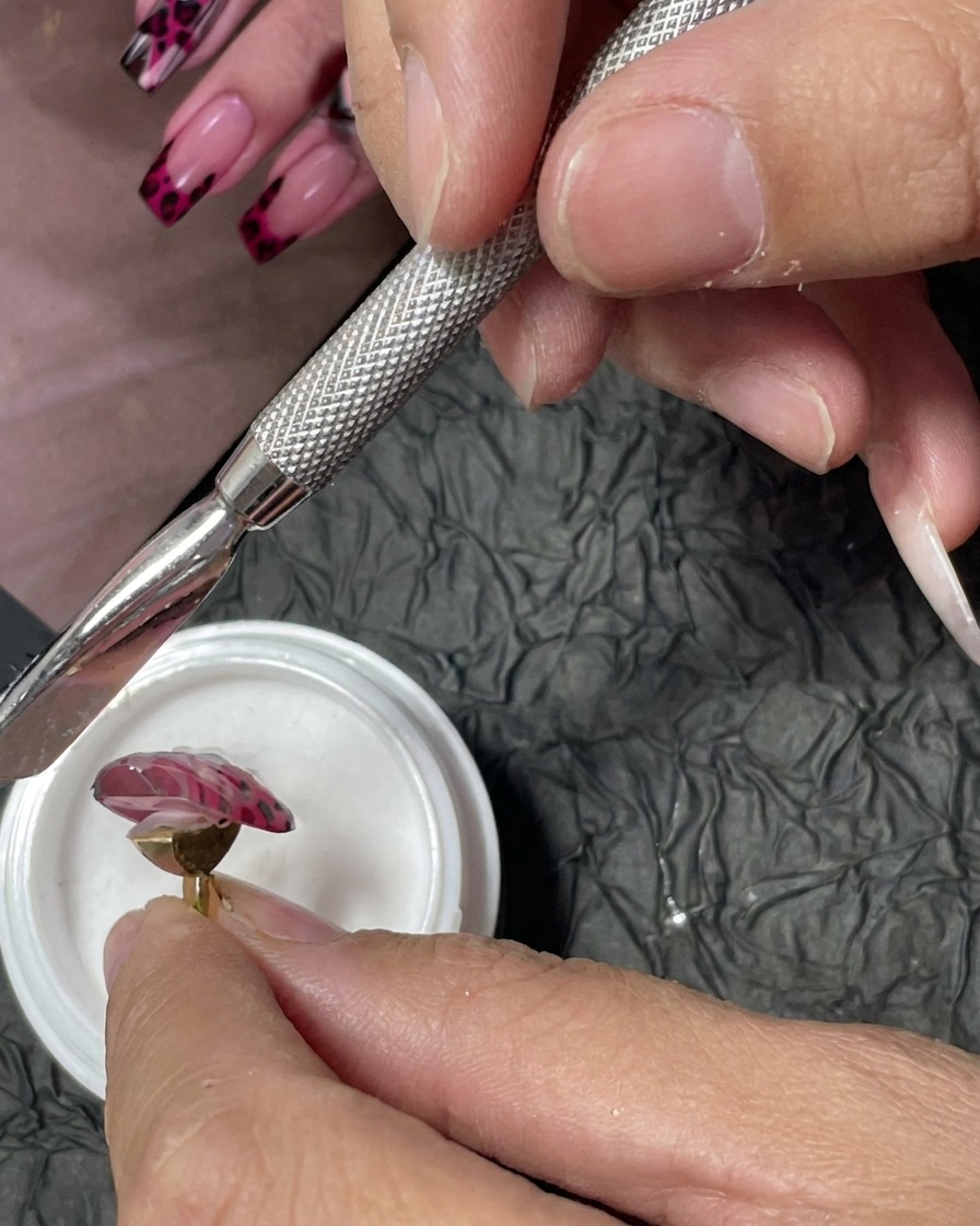 Ein Maniküre-Werkzeug wird verwendet, um feines, weißes Acrylpulver über nasse rote Gel-Punkte auf einem pinken Nagel zu streuen.