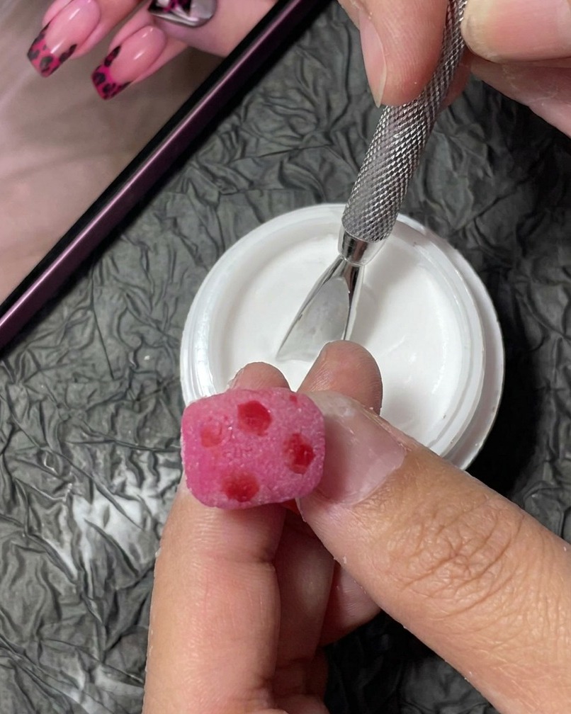 Nahaufnahme eines feinen Liner-Pinsels, der kleine, unregelmäßige schwarze Flecken auf einen pinken, texturierten Nagel malt.