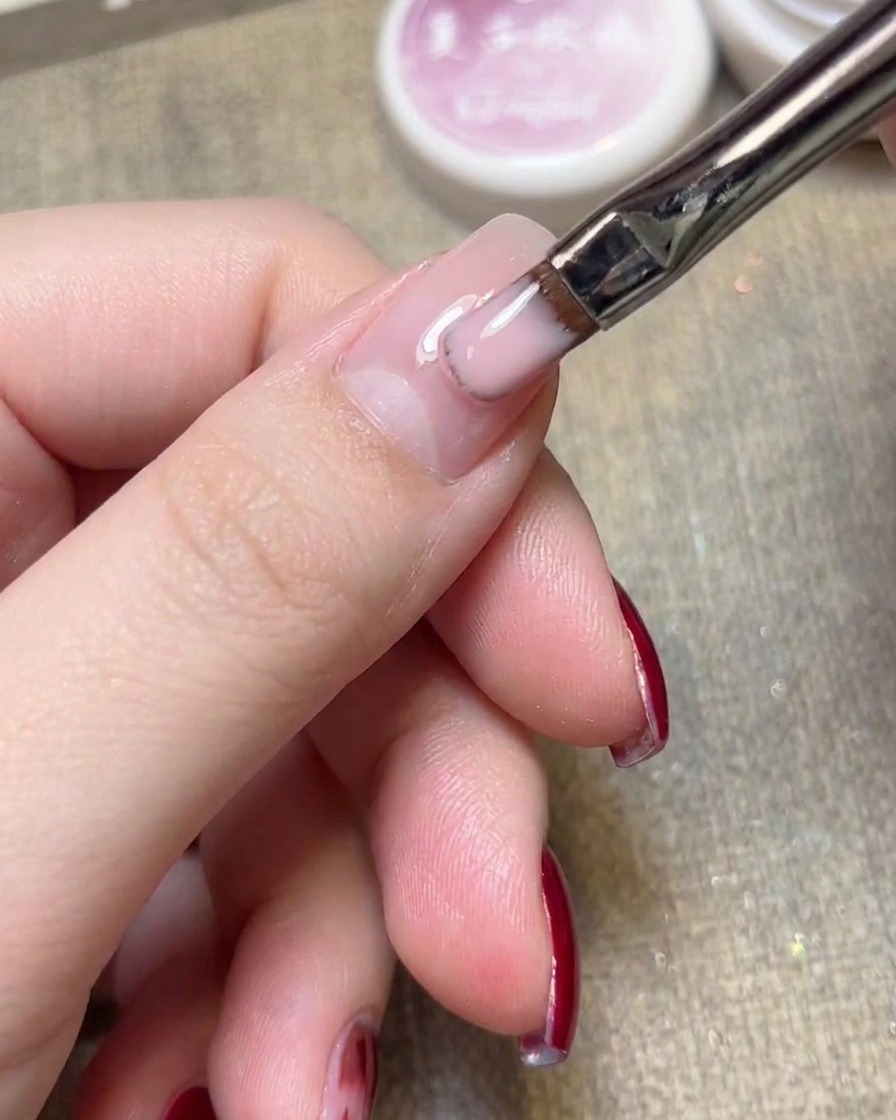 Nahaufnahme eines flachen Gel-Pinsels, der hellrosa Jelly-Nagellack auf einen Naturnagel aufträgt.