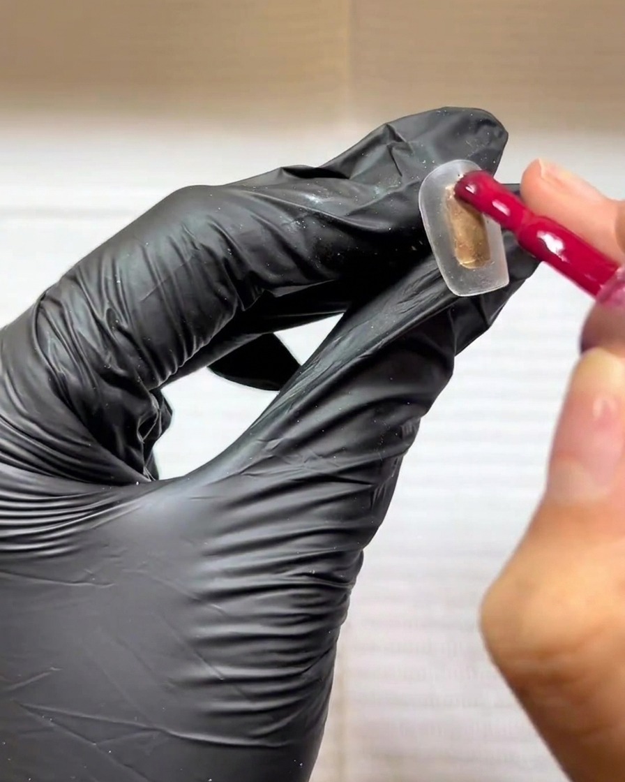 Ein behandschuhter Finger trägt mit einem Gelpinsel eine rosarote Farbe auf einen transparenten Nageltip auf.