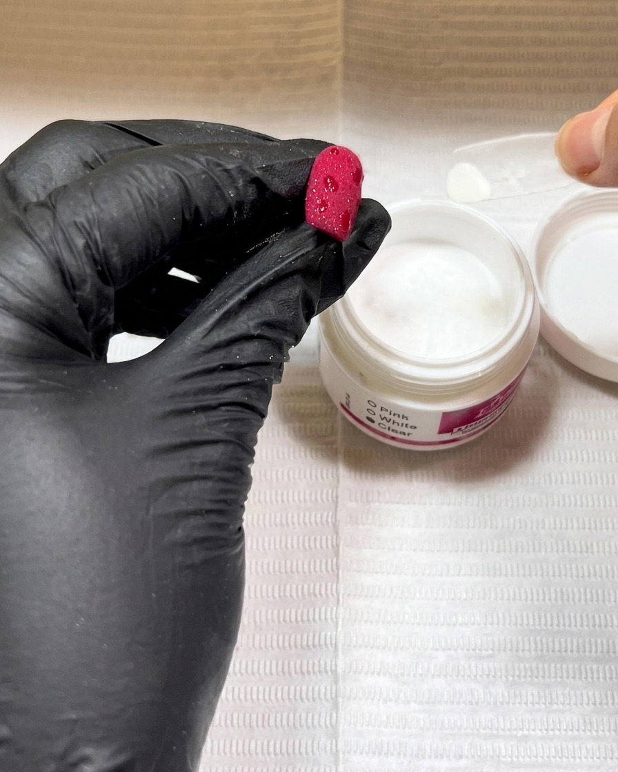 Behandschuhte Hände halten einen roten Nagel über ein Glas mit klarem Pulver und streuen das Pulver auf nasse Gelflecken.