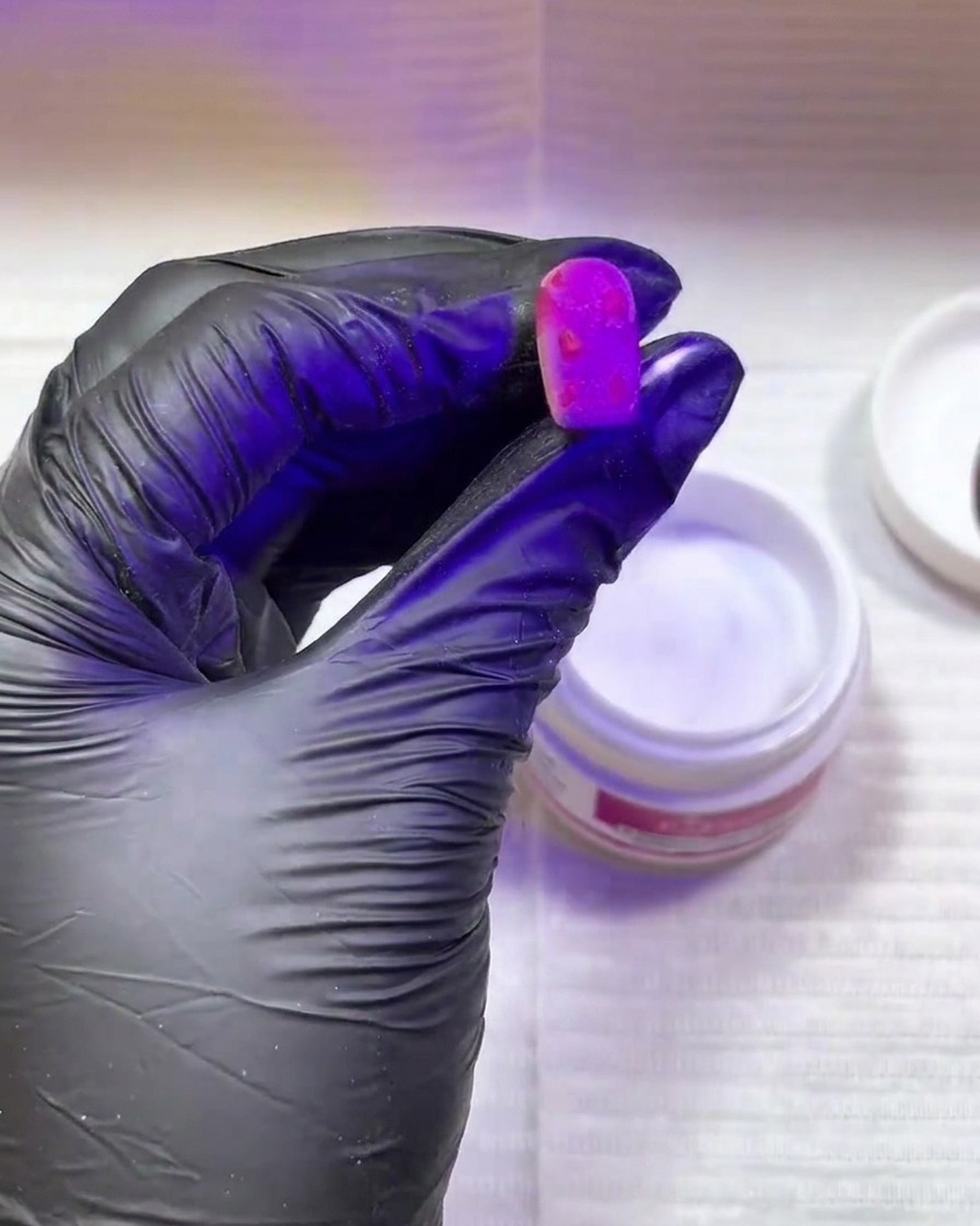 Eine Hand mit einem schwarzen Handschuh hält einen leuchtend pinken Nagel mit strukturierten Flecken unter eine violette UV/LED-Härtungslampe.