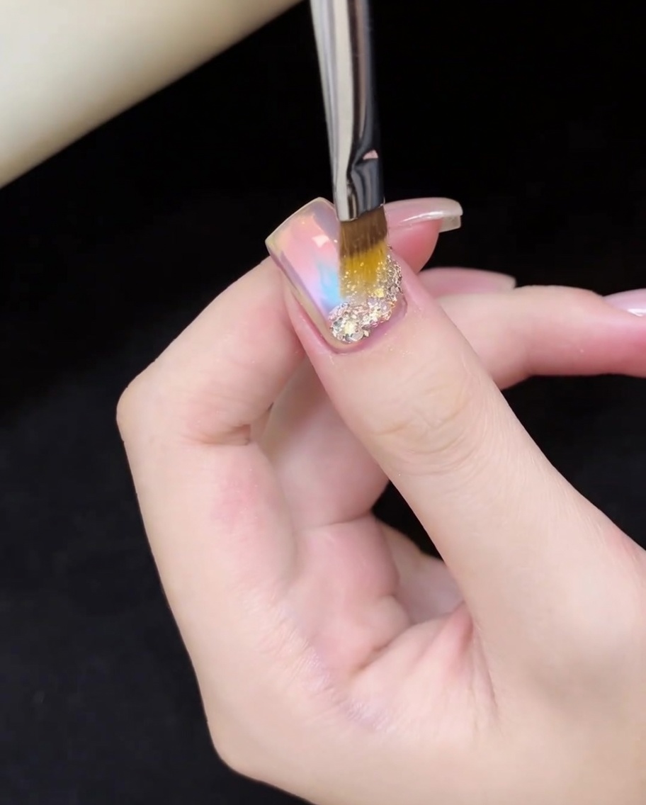 Ein Nagelpinsel platziert präzise schimmernde, irisierende Glitzerflocken auf der Nagelbasis.