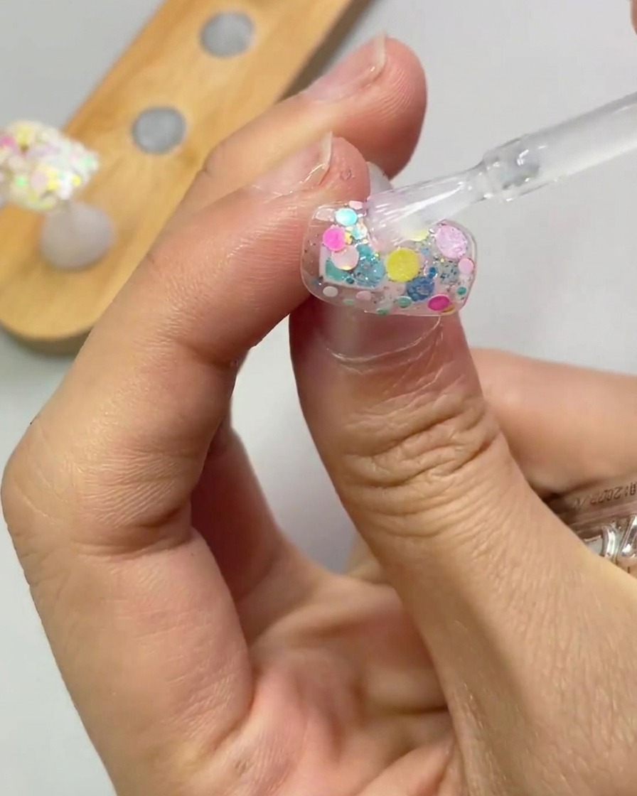 Ein Pinsel trägt eine klare Schicht Verstärkungsgel über den glitzernden Nagel auf.