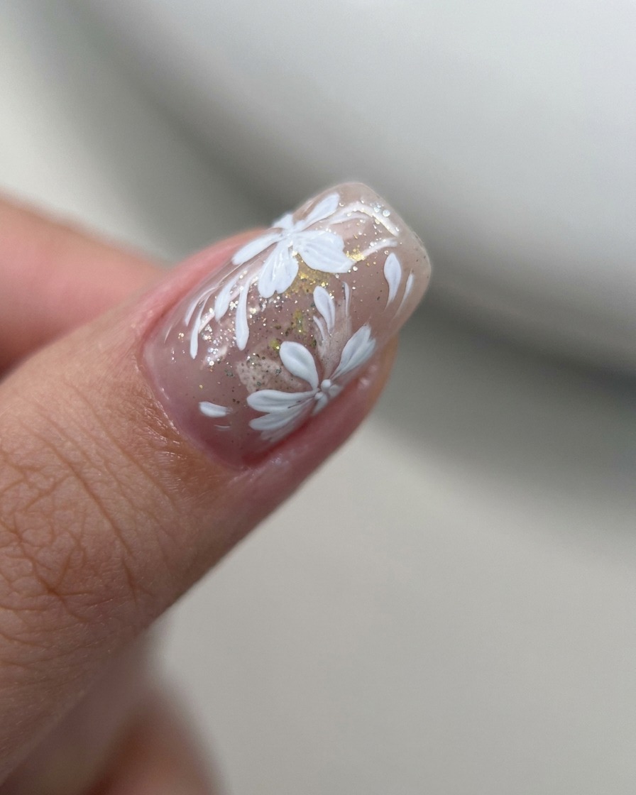 Nahaufnahme eines Liner-Pinsels, der kleine weiße Blütenblätter zu einem bestehenden Blumendesign auf einem Nude-Nagel hinzufügt.