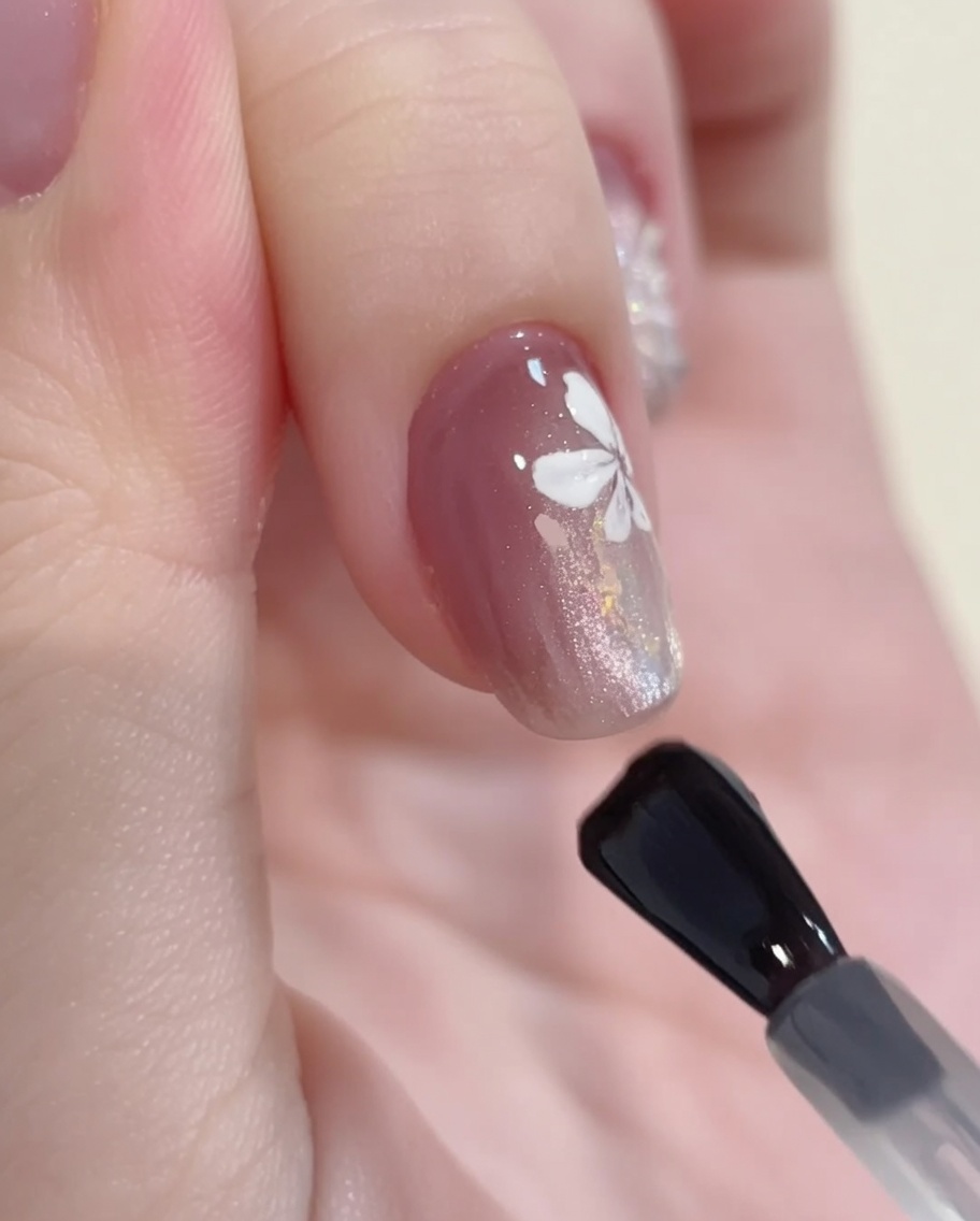 Ein Gelpinsel trägt einen klaren, glänzenden Top Coat über ein fertiges Nageldesign mit Cat-Eye-Gel, Glitzer und weißer Blumenmalerei auf.