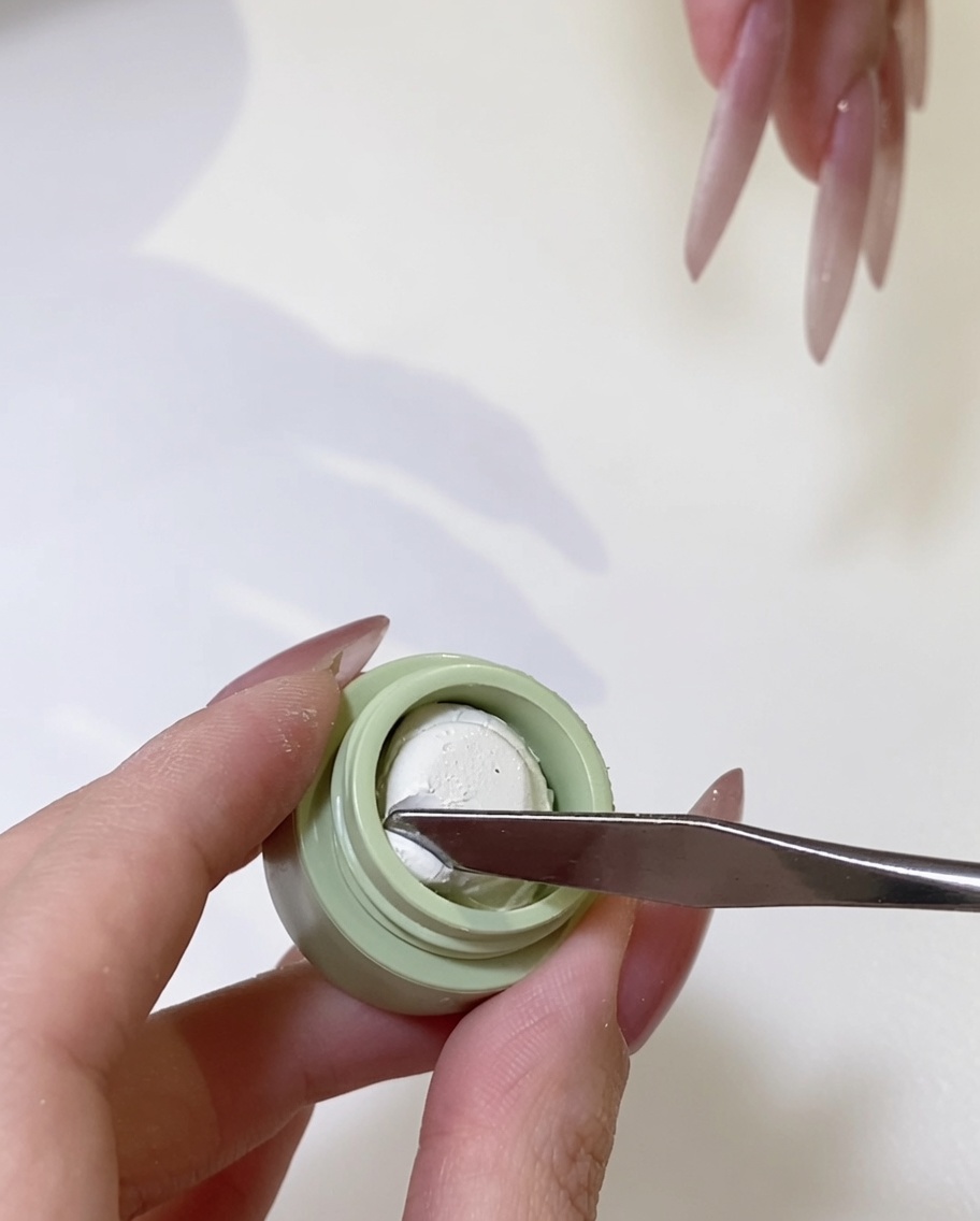 Eine Metallspachtel nimmt eine kleine Menge elfenbeinweißes Gel aus einem Tiegel auf.
