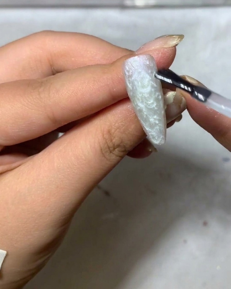 Ein feiner Liner-Pinsel beginnt damit, zarte weiße Gänseblümchen-Blütenblätter auf einen matten, grau getönten Nagel zu malen.