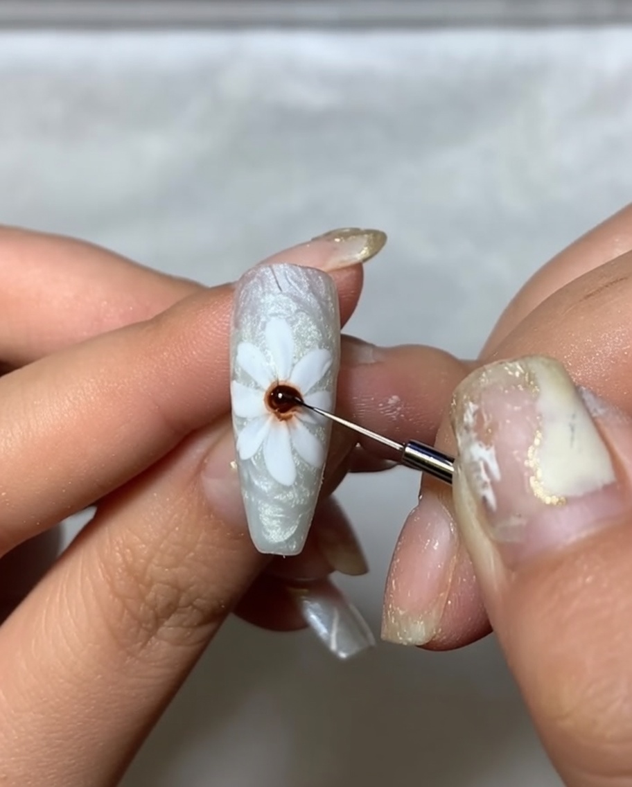 Nahaufnahme eines Nagels mit weißen Blütenblättern, wobei ein feiner Pinsel einen Punkt aus dunkelbraunem Gel in die Mitte setzt.