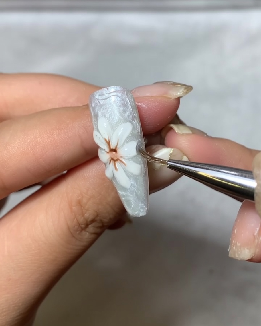Ein Applikator-Pinsel poliert vorsichtig metallisches Spiegelpuder auf die erhabenen weißen Blütenblätter des Nageldesigns.