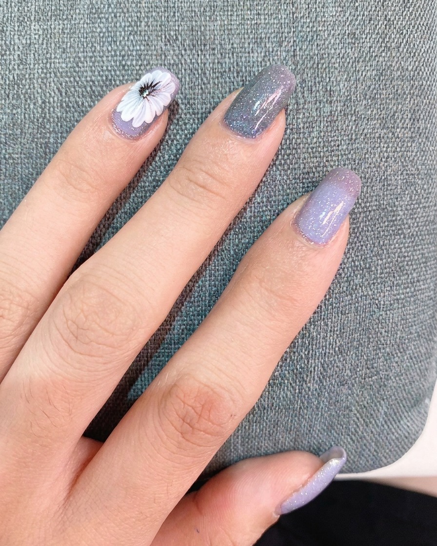 Eine Hand trägt eine dünne Schicht Top Coat über einem fertigen Nageldesign mit einem weißen Gänseblümchen und einem kleinen Text-Sticker auf.
