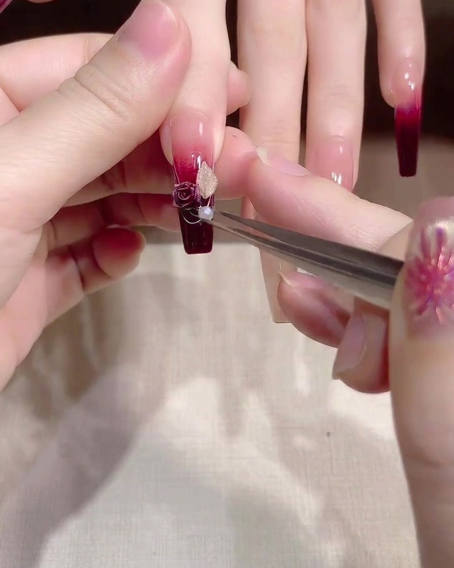Eine Pinzette setzt eine kleine, glänzende weiße Perle in die Mitte eines floralen Charms auf dem Nagel.