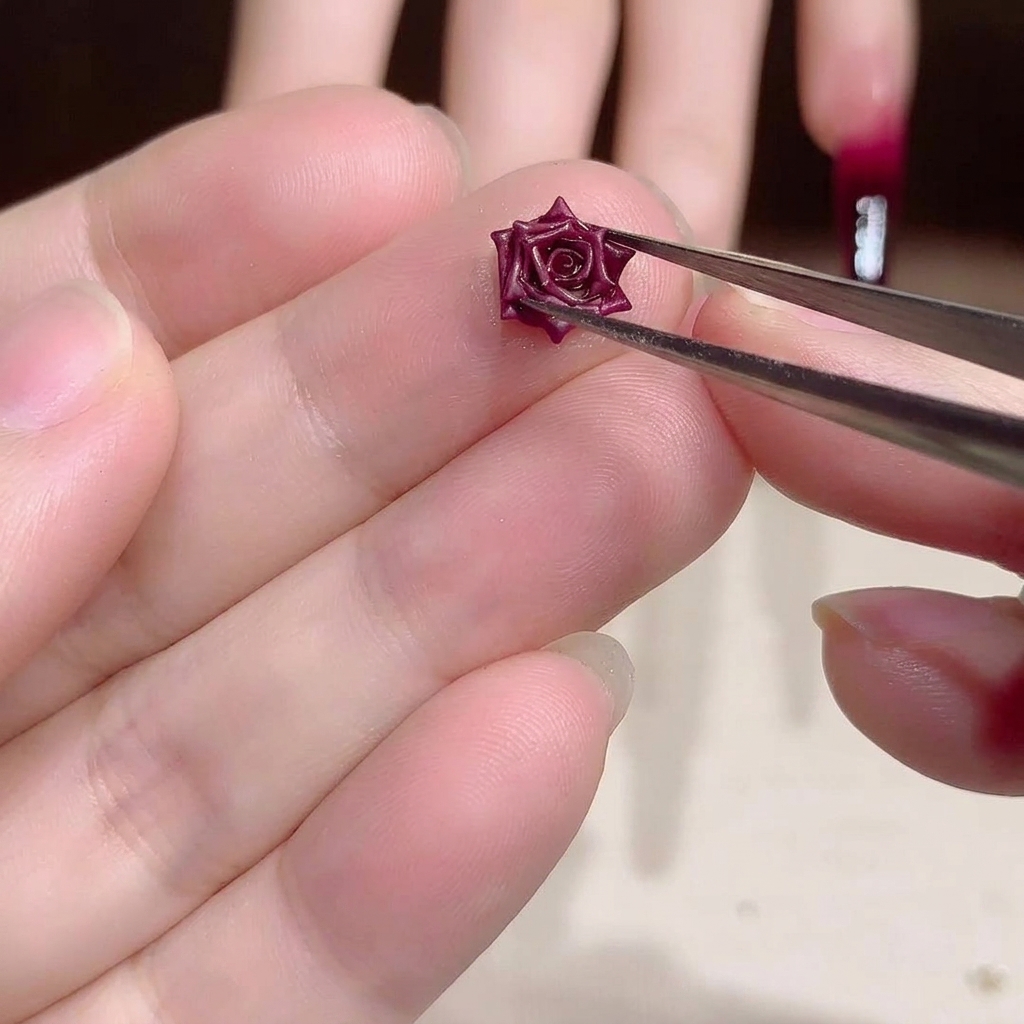 Nahaufnahme einer Metallpinzette, die ein kleines, filigranes, dunkelrotes 3D-Rosen-Charm für Nail Art hält.