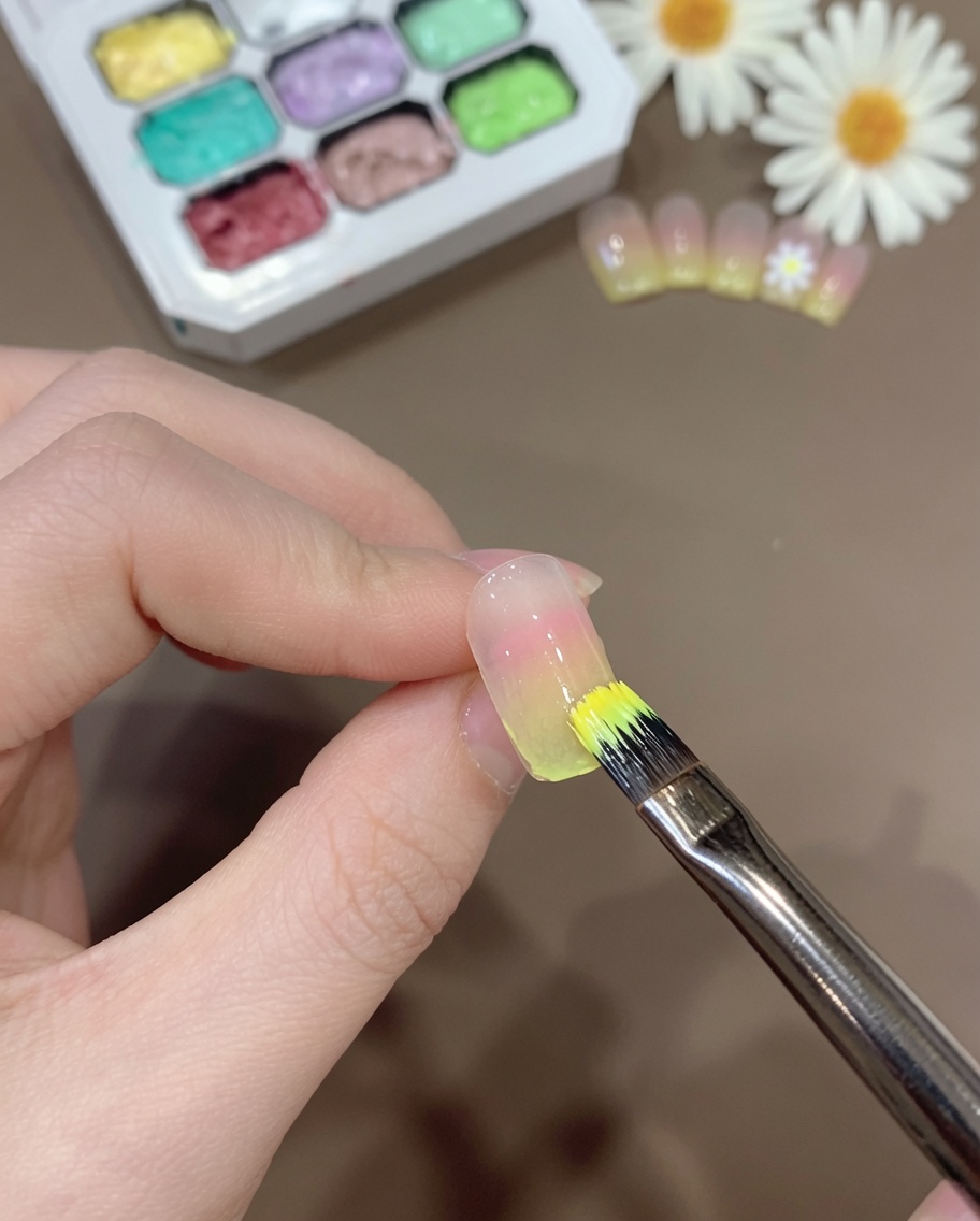 Ein Pinsel trägt eine dünne Schicht durchscheinender gelber Gelmischung auf die Spitze eines Ombré-Nagels auf.