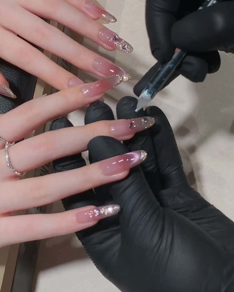 Nahaufnahme einer Nageldesignerin, die mit einem Präzisionswerkzeug einen kleinen Kristall an der Nagelhautlinie eines rosa Cat-Eye-Nagels platziert.