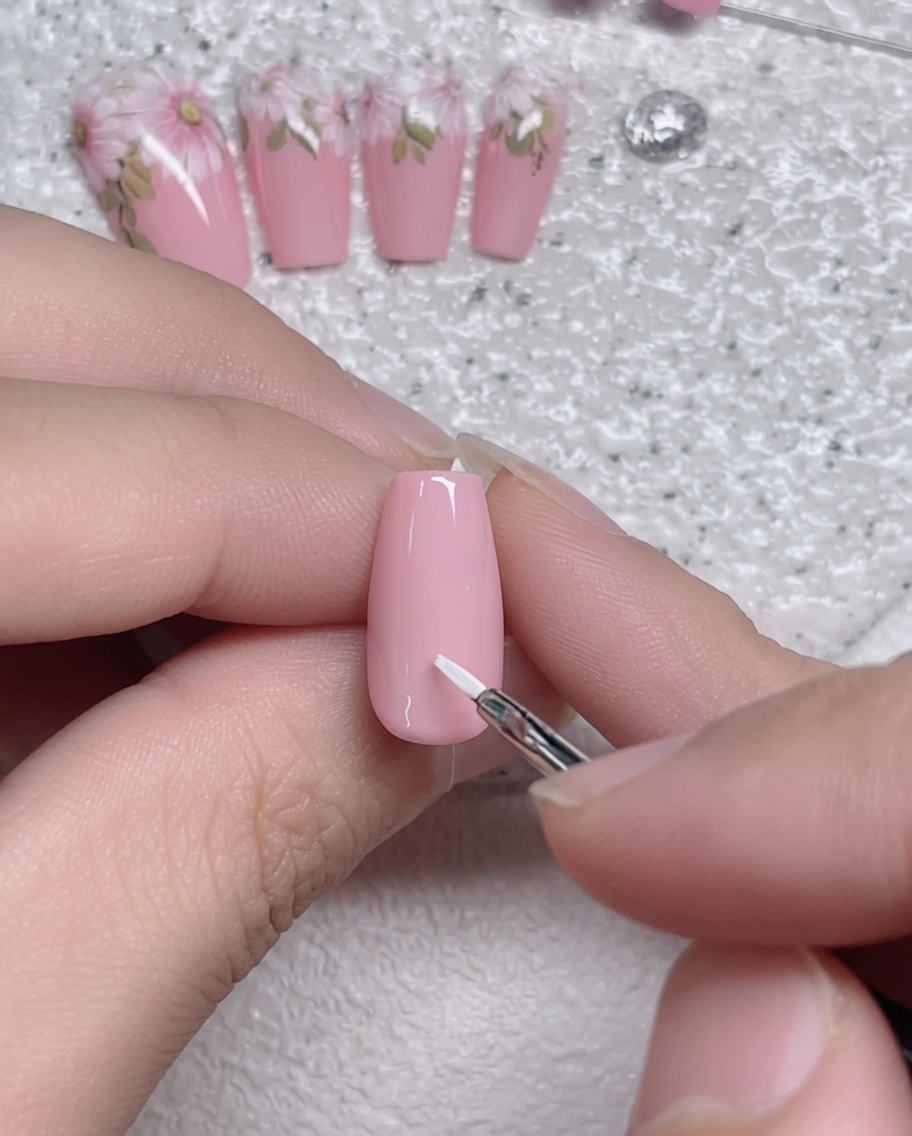 Nahaufnahme eines feinen Liner-Pinsels, der einen einzelnen weißen Blütenblattstrich auf einen rosa grundierten Nagel aufträgt.
