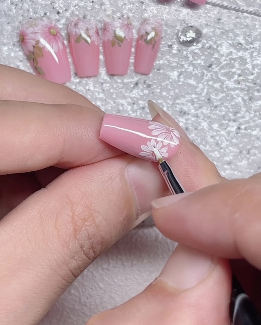 Ein feiner Liner-Pinsel, der sorgfältig die Umrisse der Gänseblümchen-Blütenblätter auf einem rosa Nagel nachzeichnet.