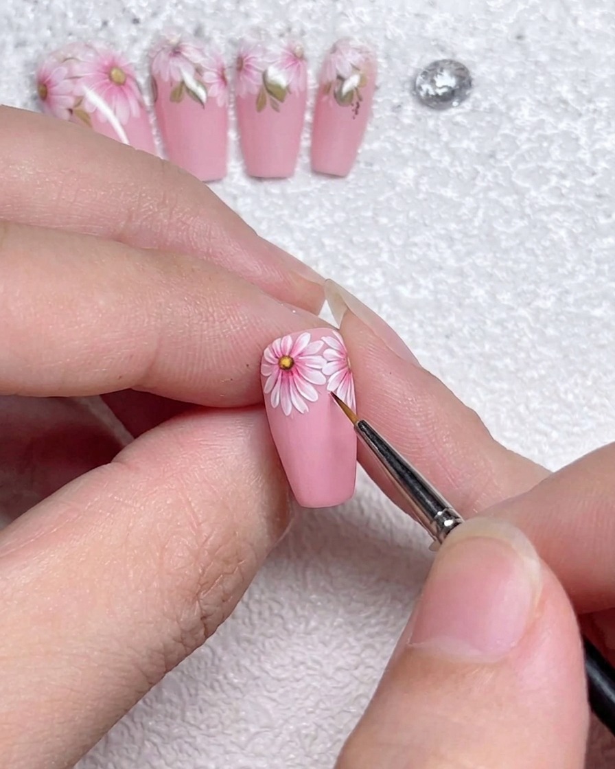 Nahaufnahme eines feinen Liner-Pinsels, der präzise weiße Highlight-Linien auf die Kanten der Gänseblümchen-Blütenblätter auf einem rosa Nagel aufträgt.