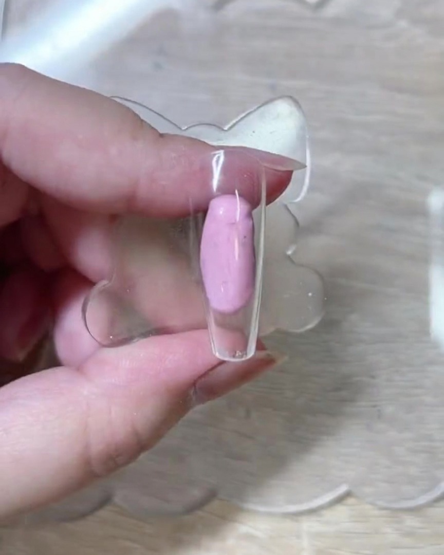 Eine Nahaufnahme zeigt einen Fine-Liner-Pinsel, der filigrane weiße Rosenkonturen auf eine unlackierte, transparente rosa Nagelspitze zeichnet.