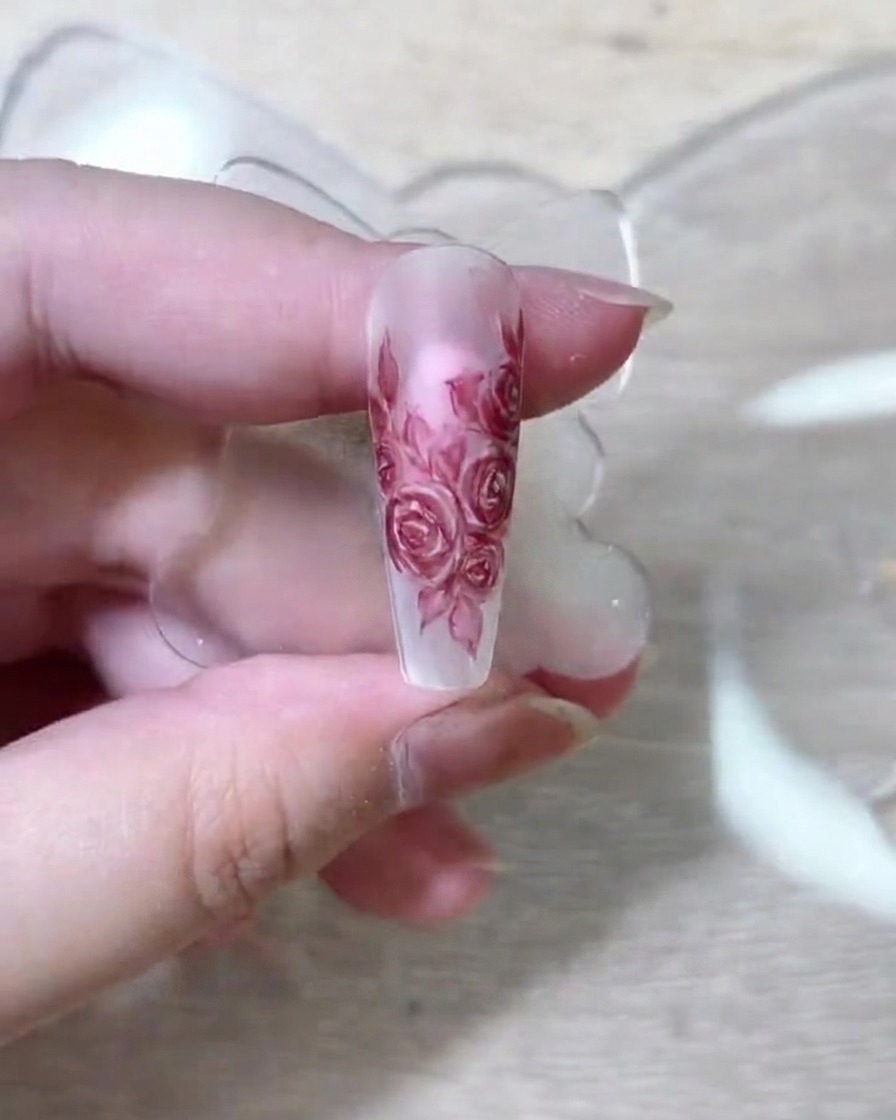 Ein feiner Nail-Art-Pinsel gibt tiefrotes Pigment auf die geschichteten Rosenblüten einer Nude-Nagelverlängerung ab.
