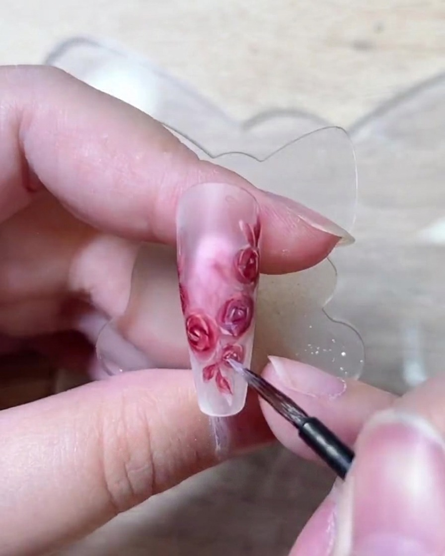 Eine Nahaufnahme eines feinen Nagelpinsels setzt filigrane weiße Highlight-Punkte auf den Rand bemalter Rosenblüten.