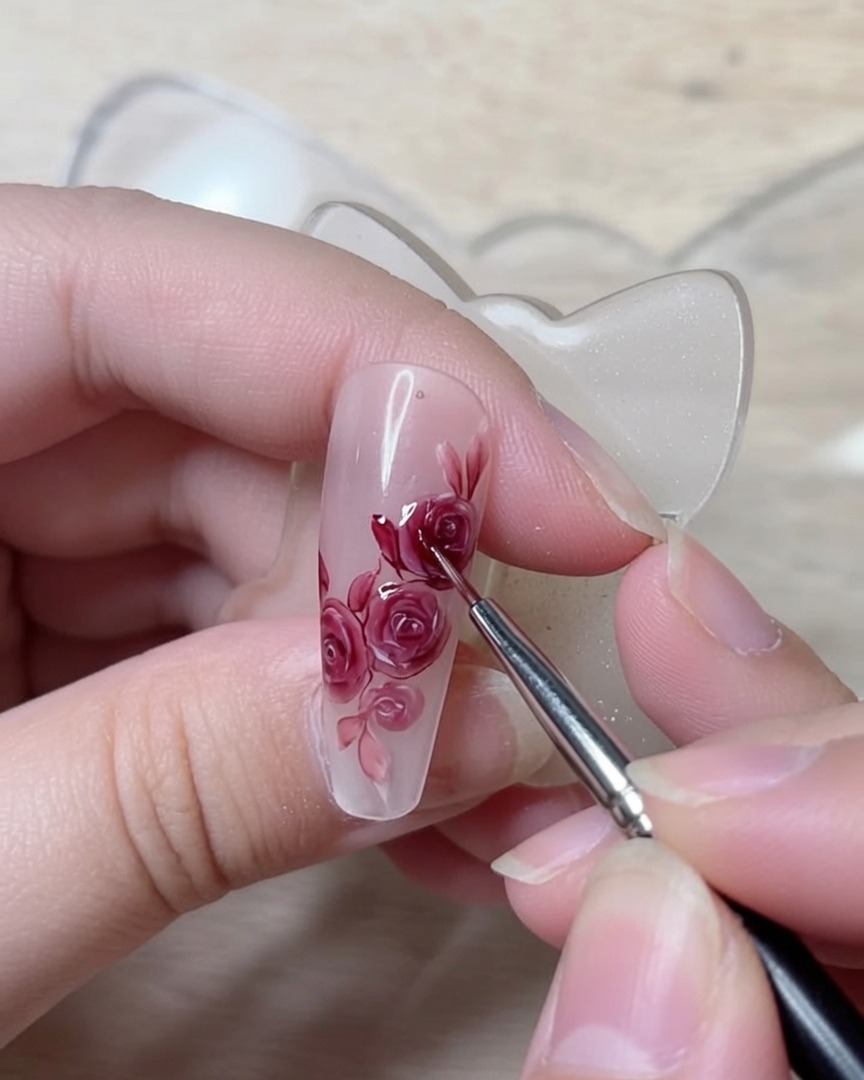 Ein feiner Detailpinsel zieht präzise dunkelrote Linien, um die strukturellen Blütenblattfalten eines Rosen-Nageldesigns zu definieren.