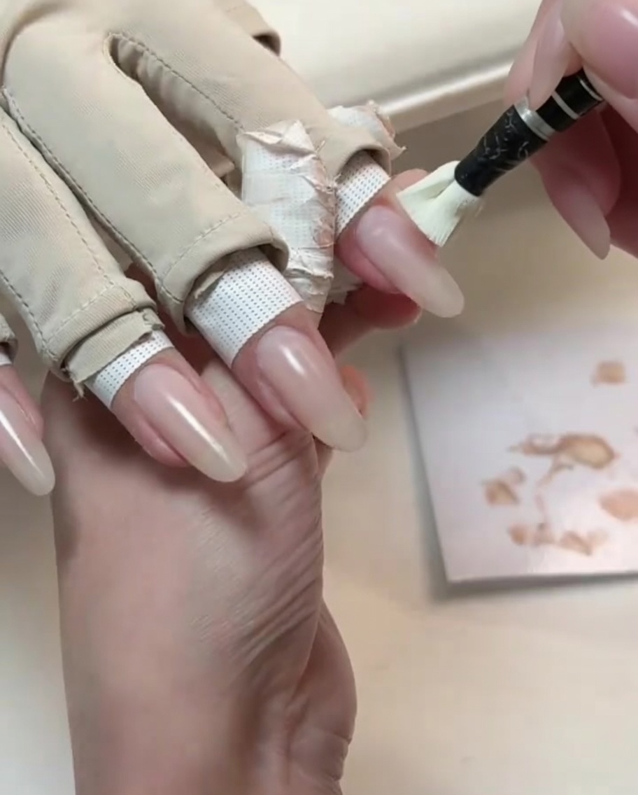 Ein kleiner Pinsel trägt einen hellen Nude-Gel-Lack auf einen langen, naturgeformten Nagel auf.