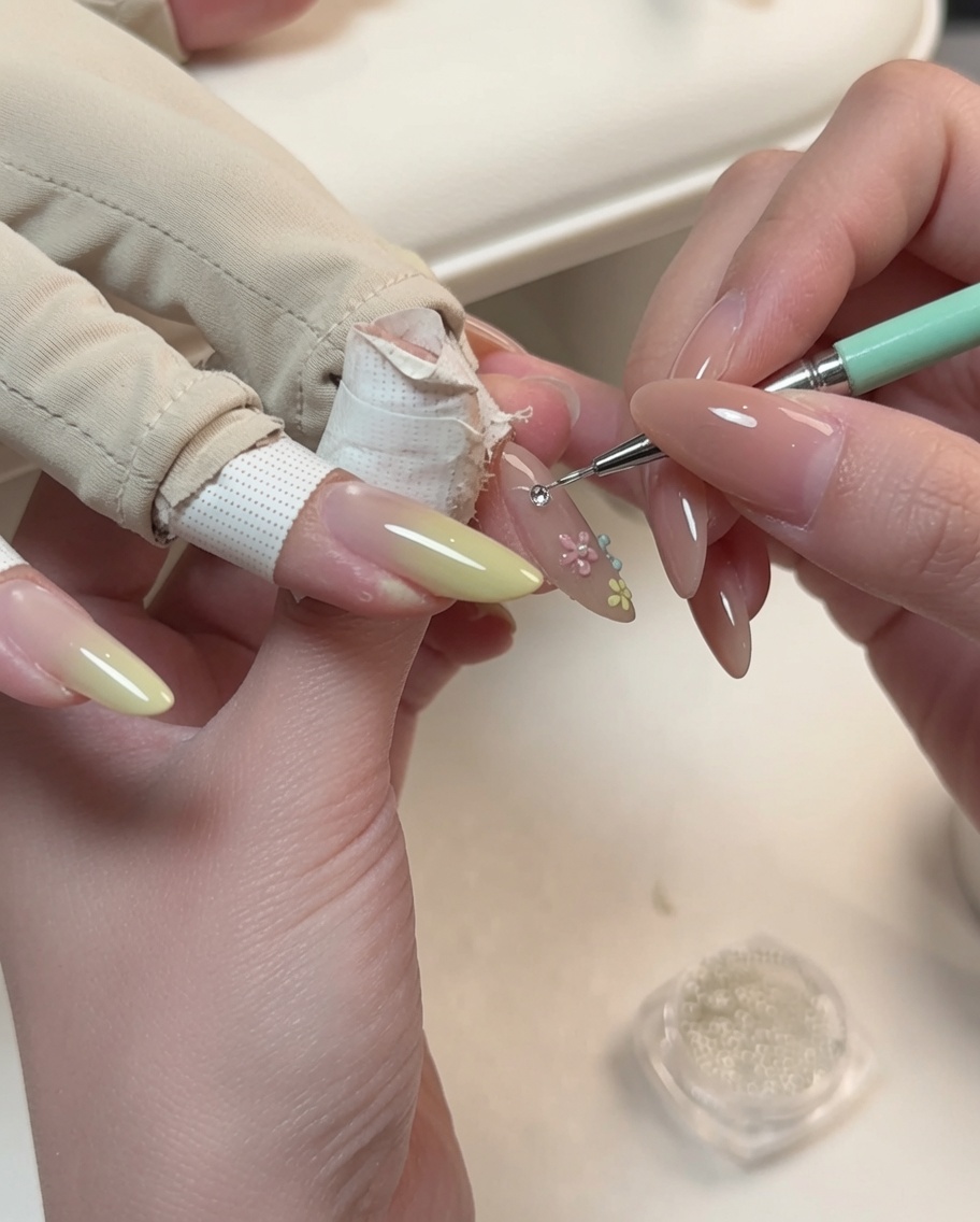 Ein Detailwerkzeug platziert eine kleine Perle in die Mitte eines floralen Nageldesigns auf einem mandelförmigen Nagel.