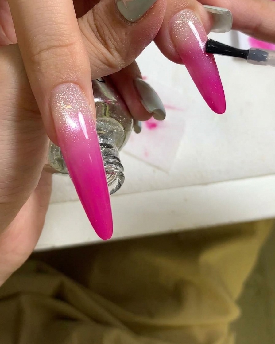 Nahaufnahme, wie ein Pinsel einen Topcoat auf einen fertigen pinken Verlaufnagel aufträgt.