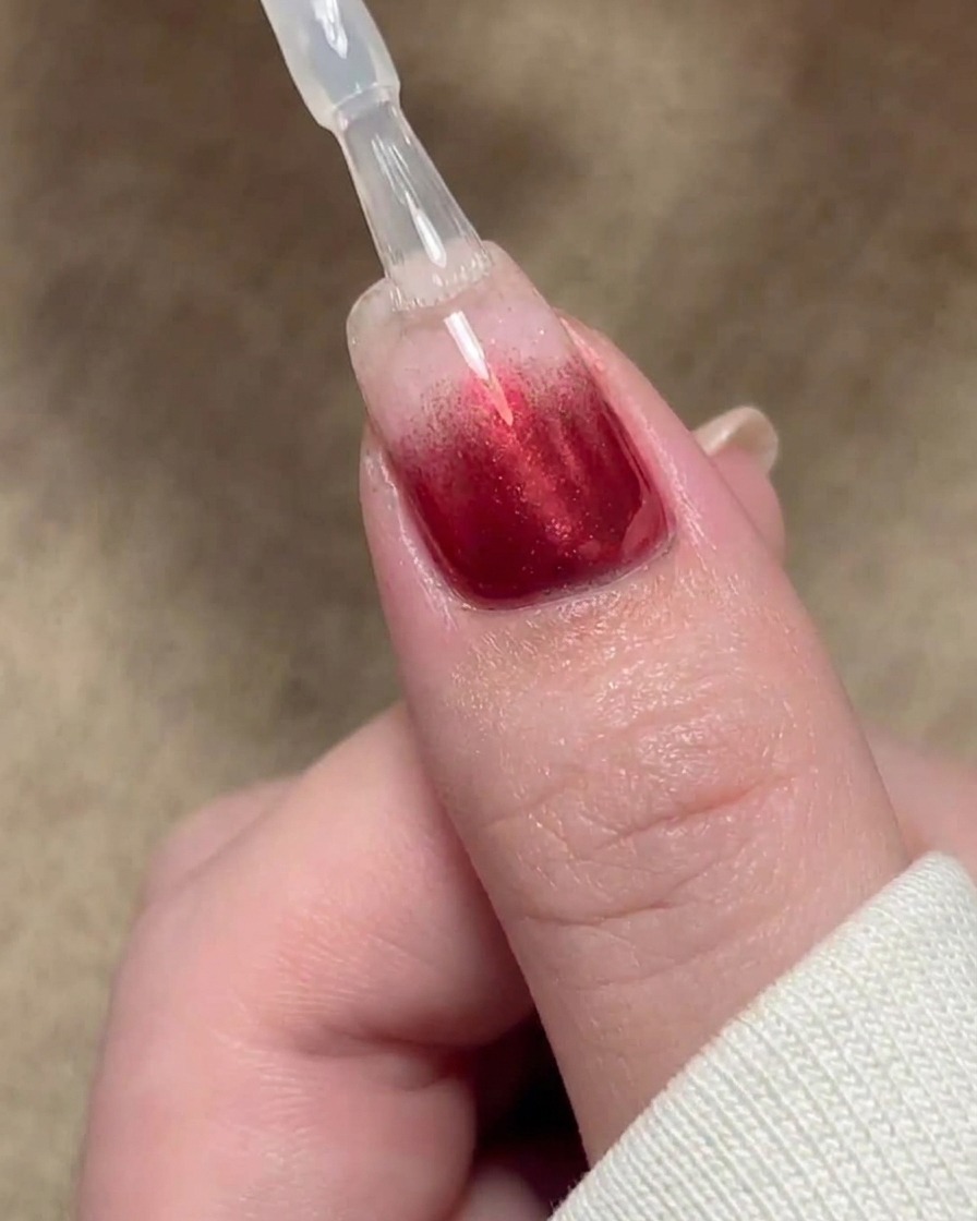 Ein Pinsel trägt eine klare Versiegelungsschicht aus Basisgel über ein rotes Gradient-Nageldesign auf.