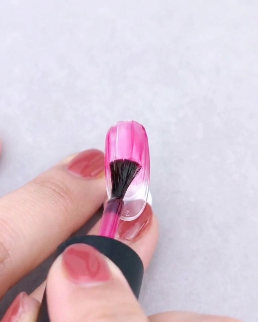 Ein Fächerpinsel, der ein rosa Gel sanft nach unten auf einem klaren Nagel verblendet, um einen Ombré-Effekt zu erzeugen.