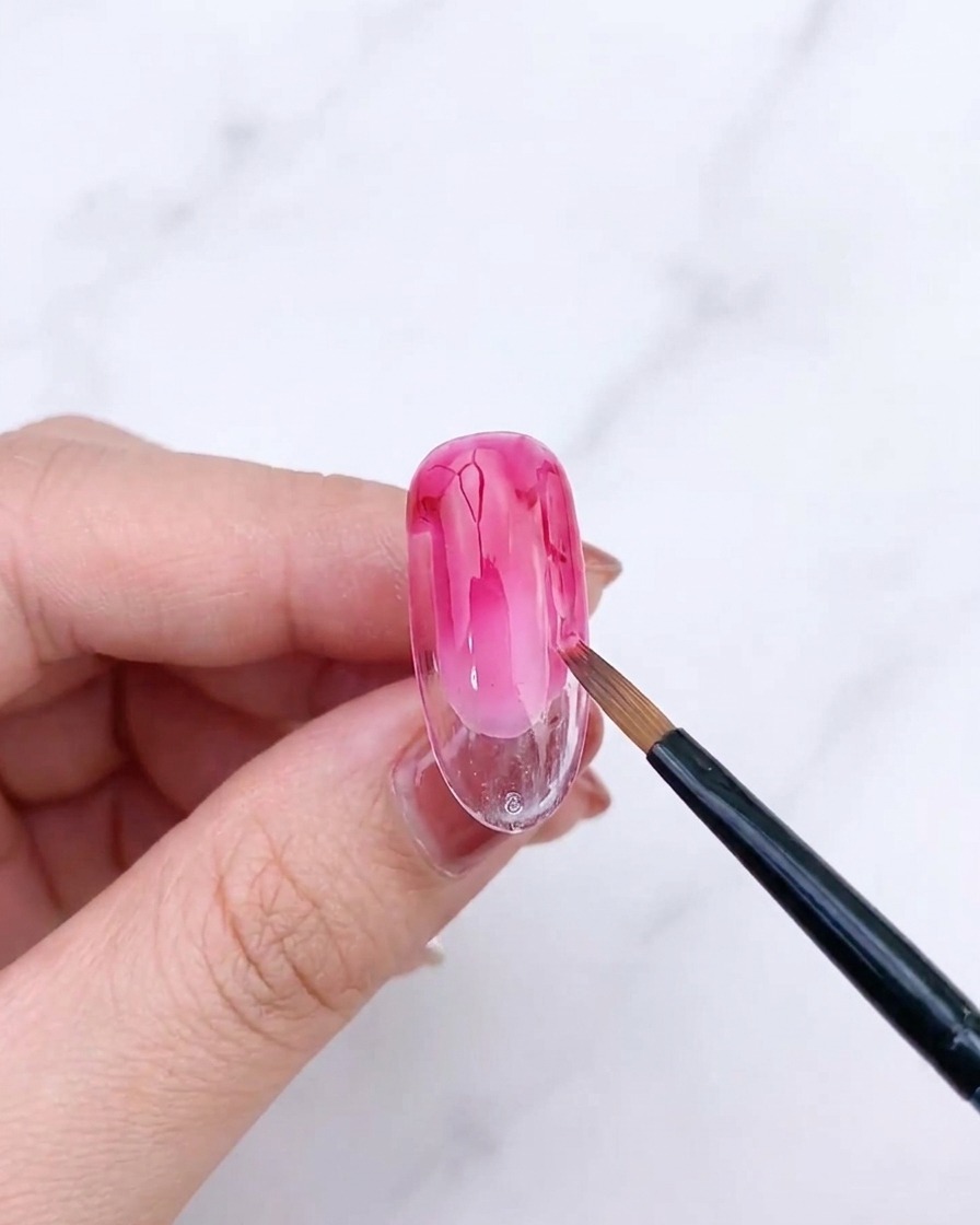 Ein Fächerpinsel verblendet die zweite Schicht rosa Gel auf einem Nagel und erzeugt einen verfeinerten Farbverlauf.