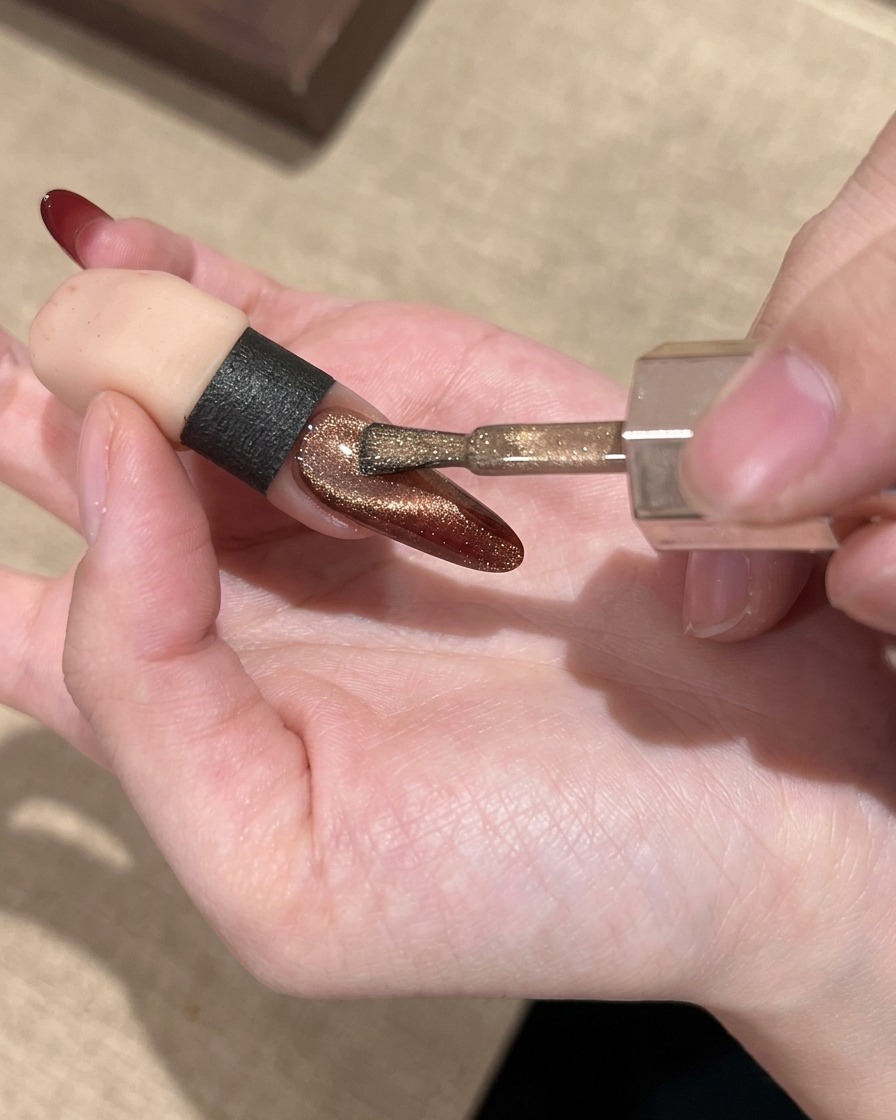 Ein Nagelpinsel trägt ein schimmerndes goldenes Cat-Eye-Gel über den rot verlaufenden Nagel auf.