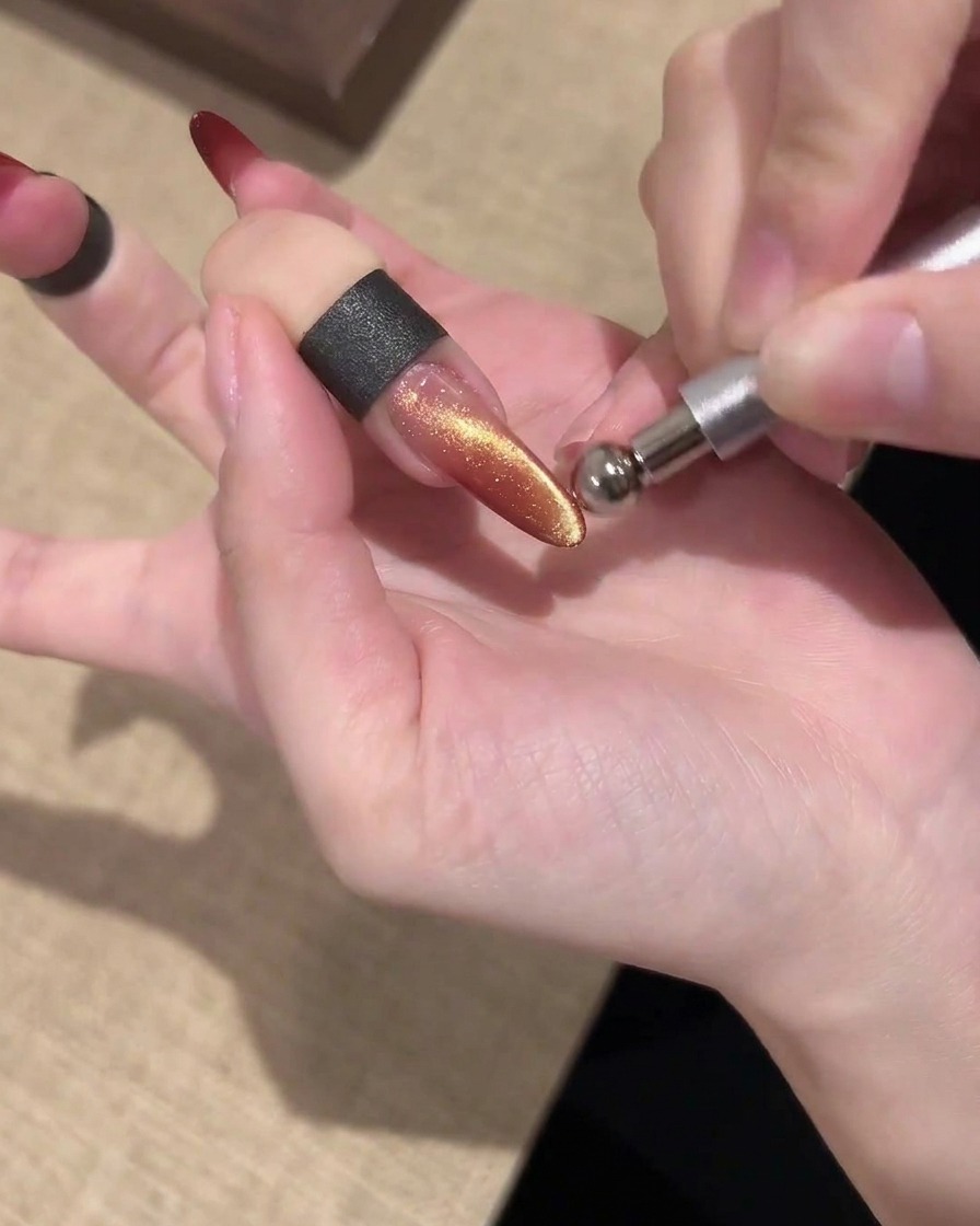 Ein Stabmagnet, der parallel über einem schimmernden Gold- und Rot-Cat-Eye-Nagel gehalten wird, um das Licht zu formen.