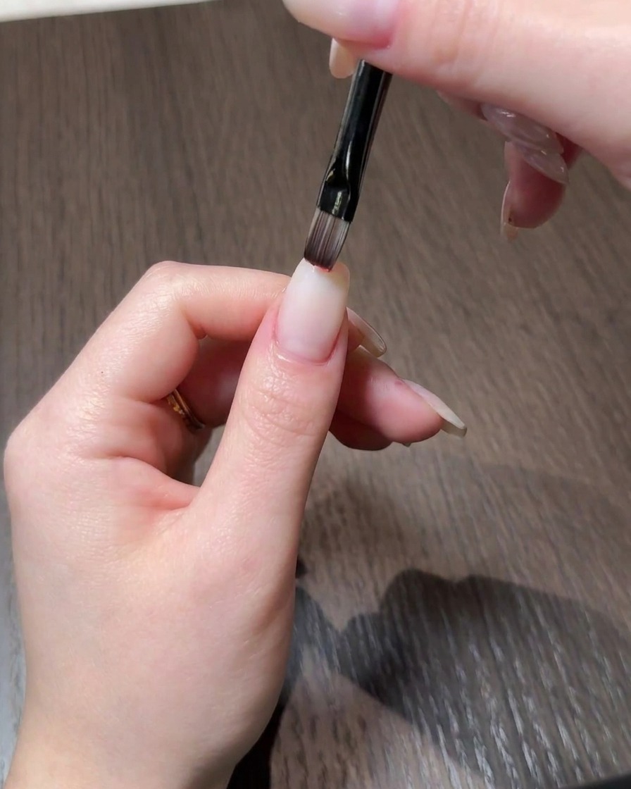 Eine Nahaufnahme einer Hand, die mit einem feinen Pinsel rotes Gel-Polish auf die Spitze eines naturnah wirkenden Nagels aufträgt.