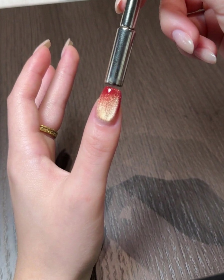 Ein Magnetwerkzeug, das Cat-Eye-Partikel zum hinteren Rand eines Rot-Gold-Verlaufsnagels schiebt.