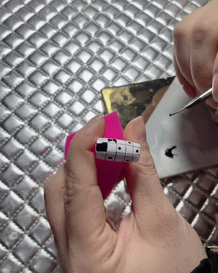 Eine Nahaufnahme einer Nageldesignerin, die einen Feinliner-Pinsel verwendet, um die Kante eines schwarzen Quadrats auf einem Schachbrett-Nageldesign zu begradigen.