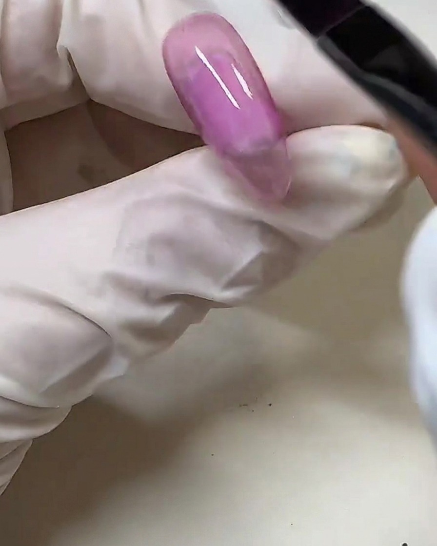Eine behandschuhte Hand trägt mit einem Gelpinsel halbtransparenten rosa Gellack auf einen klaren Nageltipp auf.