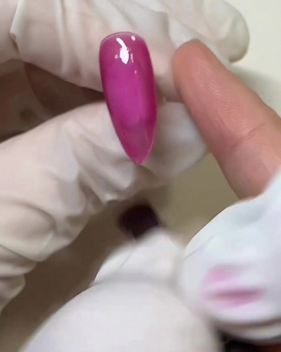Nahaufnahme eines behandschuhten Fingers, der eine dünne Schicht klares, festes Klebegel über der ausgehärteten rosa Nageloberfläche verteilt.