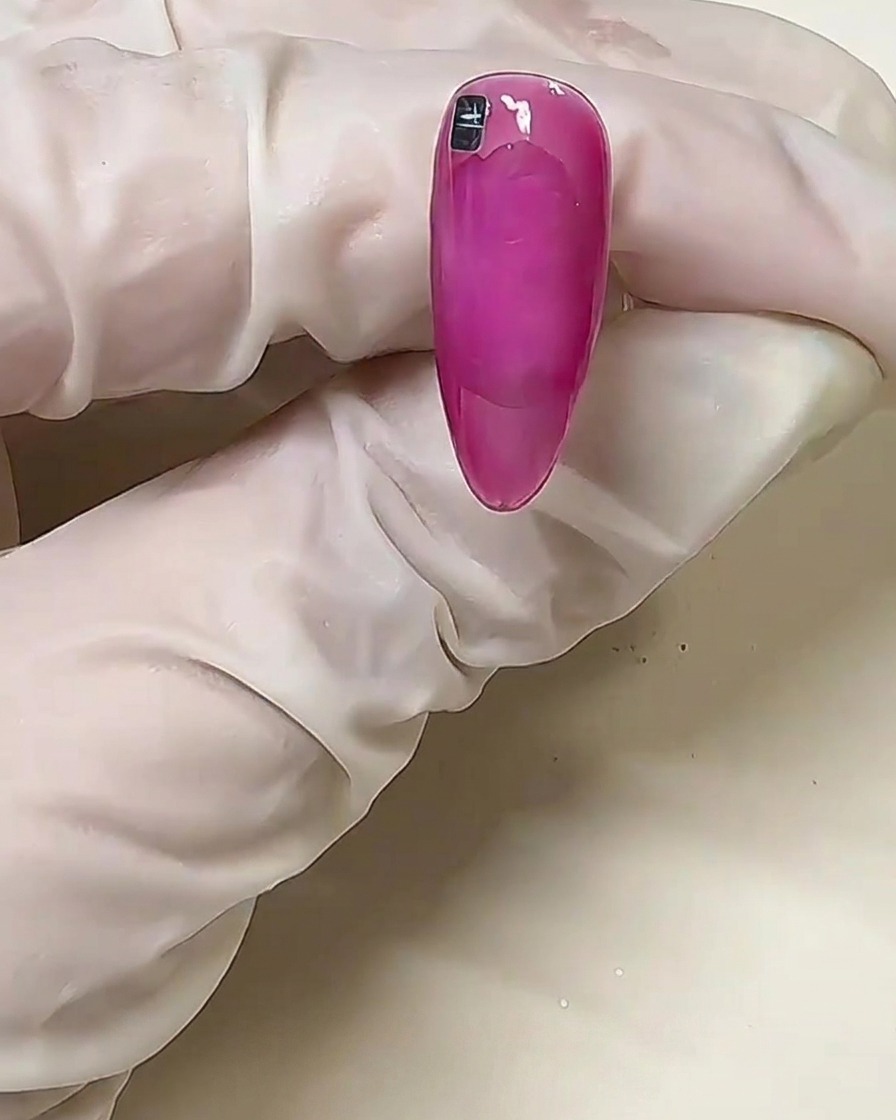 Nahaufnahme eines rosa Nagels mit einem einzelnen silbernen Metallic-Quadrat, das nahe der Nagelhaut auf dem nicht ausgehärteten festen Kleber platziert wurde.
