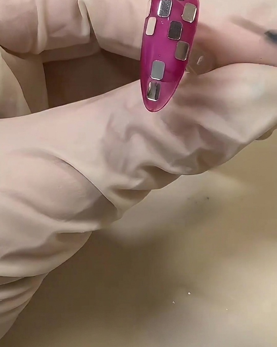 Eine behandschuhte Hand richtet kleine silberne quadratische Folienstücke auf einem durchscheinenden rosa Nagel aus, um ein Schachbrettmuster zu kreieren.