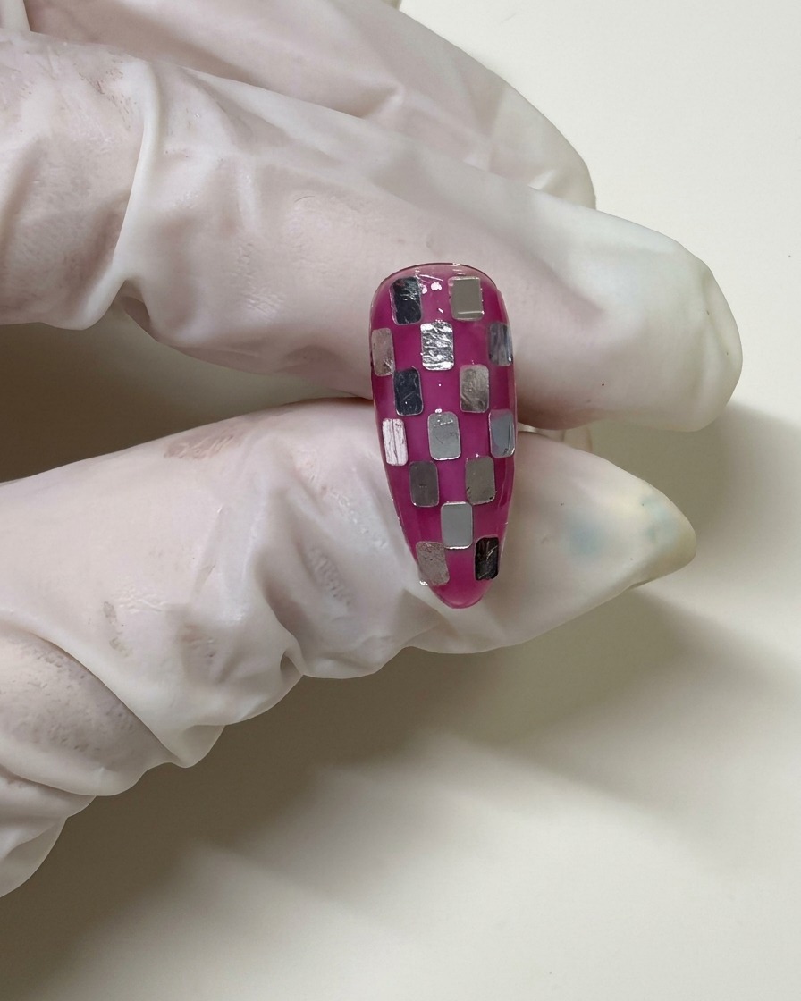 Eine behandschuhte Hand hält einen Nagel mit Schachbrettmuster, der mit einer transparenten rosa Gelschicht überzogen wurde, um die Silberquadrate zu integrieren.