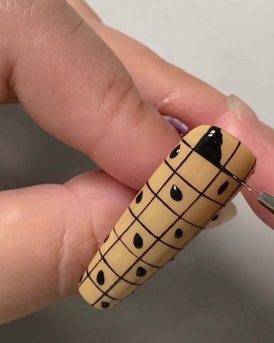 Eine Nahaufnahme eines Liner-Pinsels, der das obere linke Quadrat eines Gitters auf einem Nude-Nagel mit schwarzem Gel-Lack ausfüllt.
