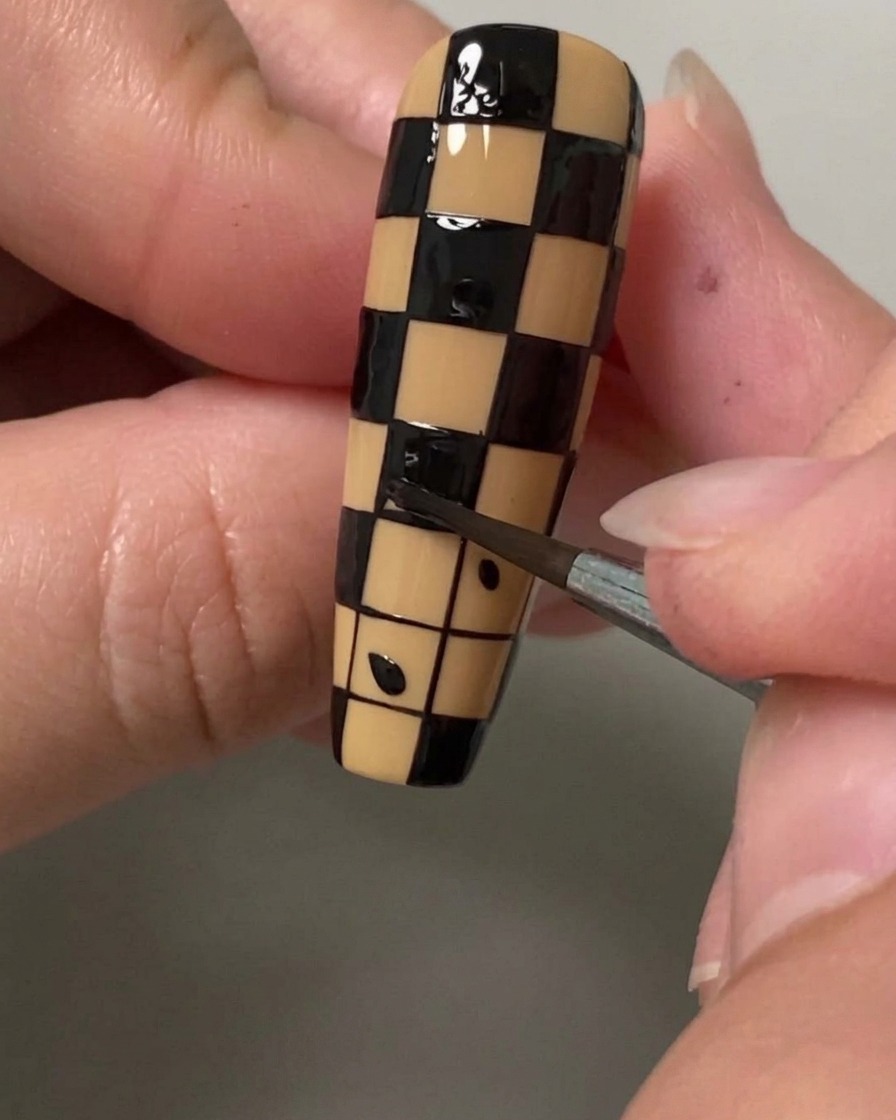 Eine Nahaufnahme eines feinen Detailpinsels, der ein kleines Quadrat mit schwarzem Lack ausfüllt, um ein Karomuster auf einem Nagel zu erzeugen.