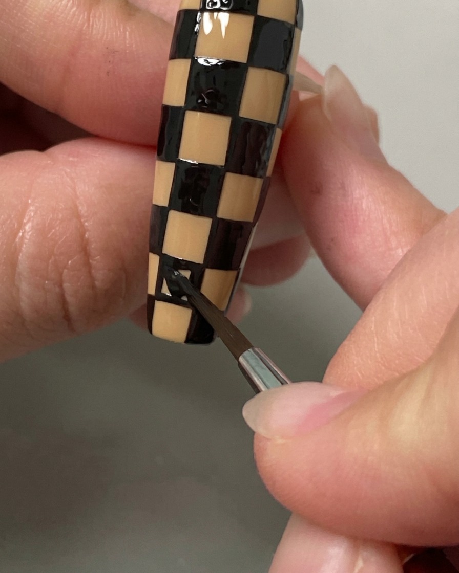 Ein Liner-Pinsel füllt die letzten Quadrate eines schwarz-nudefarbenen Karomusters auf einem langen Nagel aus.