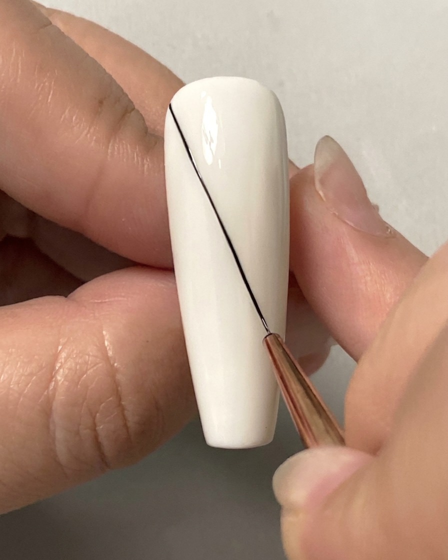 Eine Nahaufnahme eines feinen Liner-Pinsels, der eine lange diagonale schwarze Linie über einen weißen Nagel zieht.