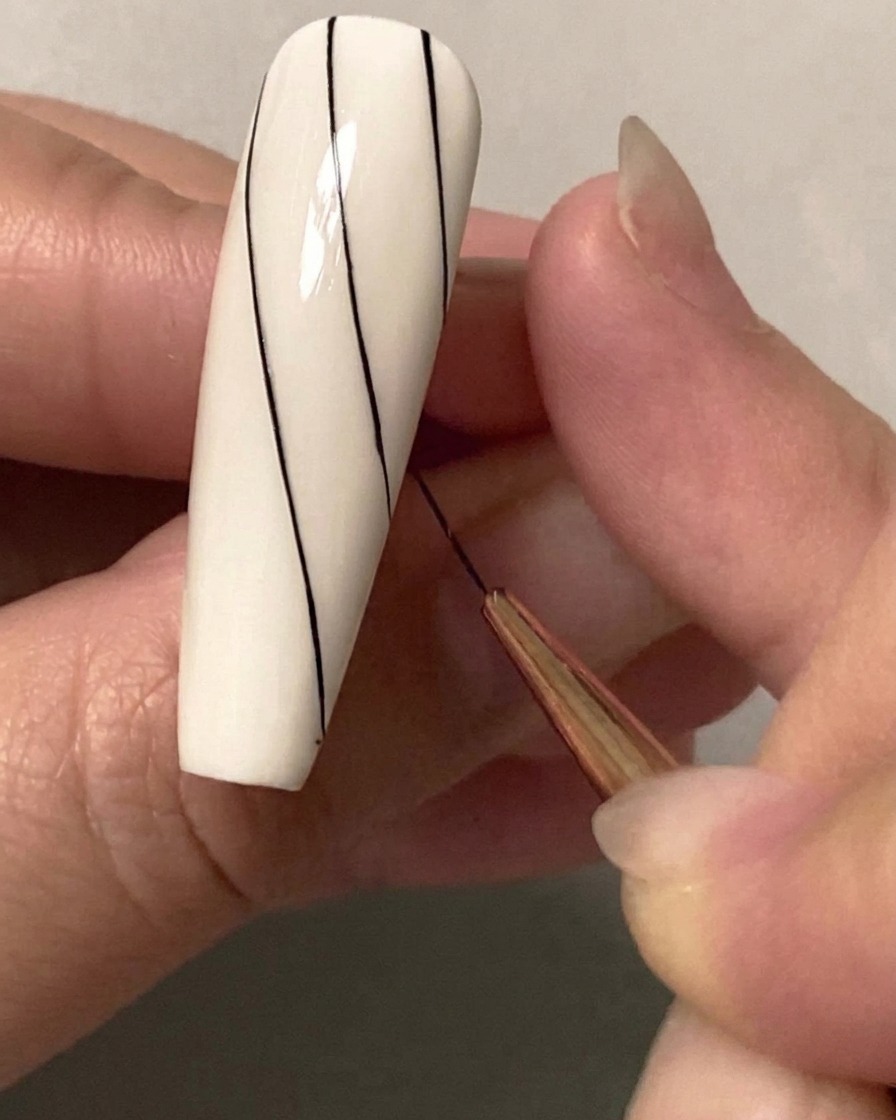 Eine Nahaufnahme eines feinen Nail-Art-Pinsels, der eine zweite parallele diagonale schwarze Linie auf der oberen Hälfte eines weißen Nagels zieht.