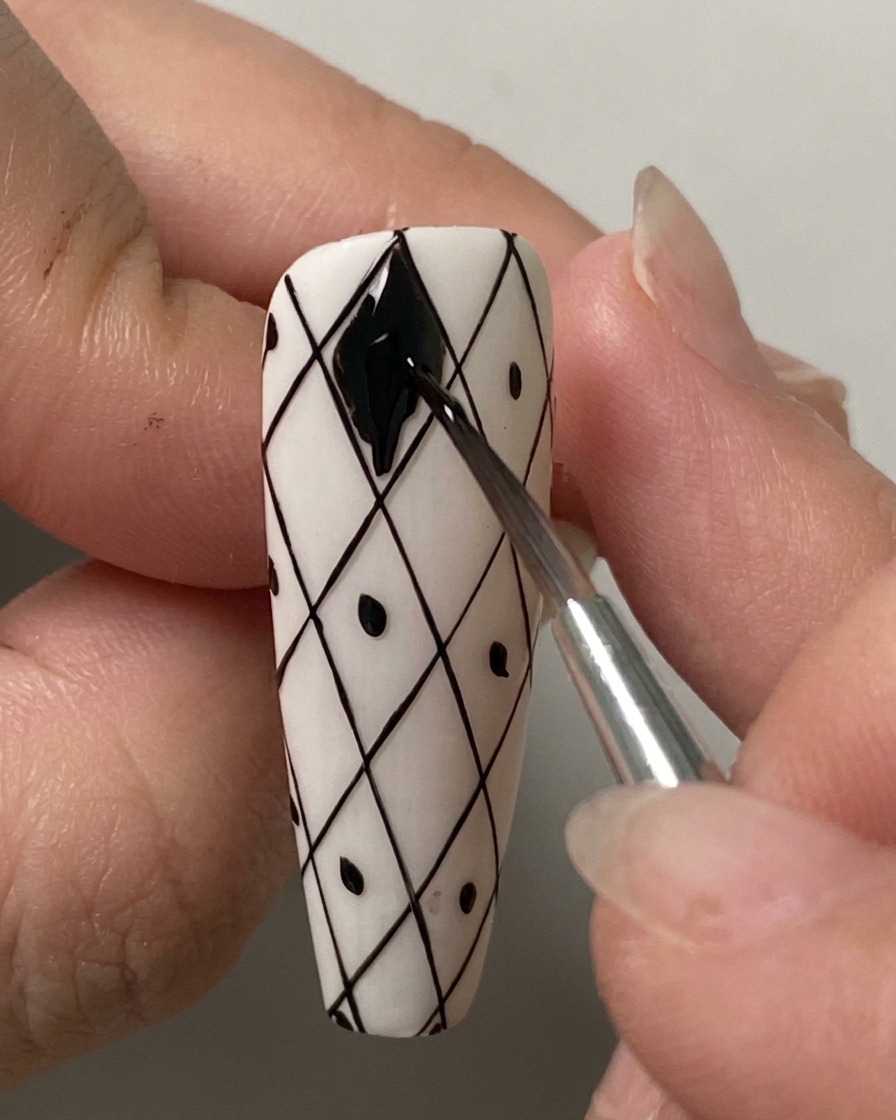 Ein feiner Liner-Pinsel füllt die oberste Rautenform auf einem vorgezeichneten weißen Gitter Nagel sorgfältig mit schwarzem Gel-Lack aus.