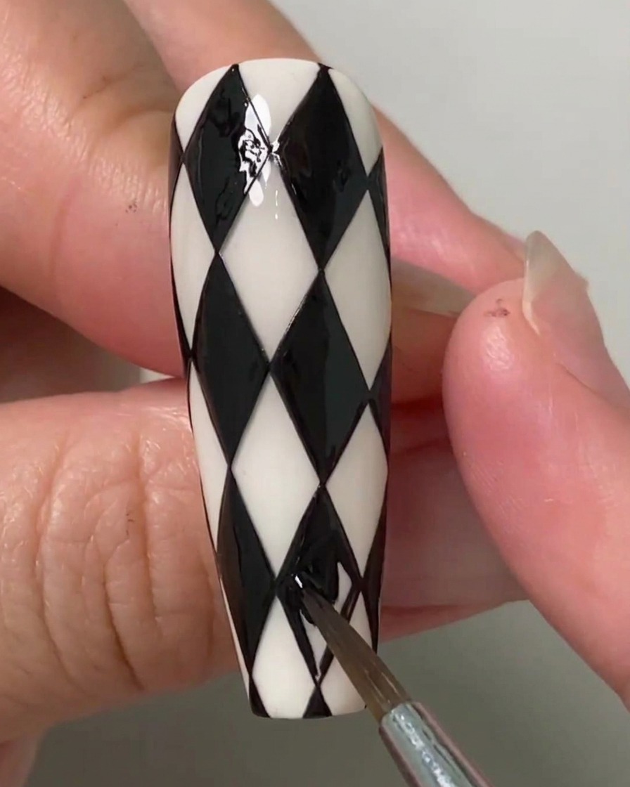 Eine Nageldesignerin verwendet einen feinen Pinsel, um die letzten verbleibenden Rautenformen auf einem schwarz-weiß gemusterten Argyle-Nagel auszufüllen.